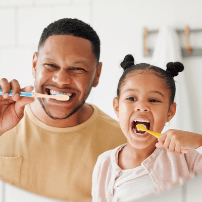 O'fallon dental works | cosmetic and general dentistry | dental care in o'fallon, mo 17 Father and daughter smiling while brushing their teeth together in the bathroom mirror, demonstrating fun ways to teach kids oral hygiene during national children’s dental health month.